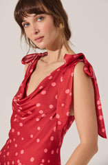 A woman wearing a sleeveless red satin dress with pink polka dots. The dress features a draped neckline and shoulder straps tied into bows.