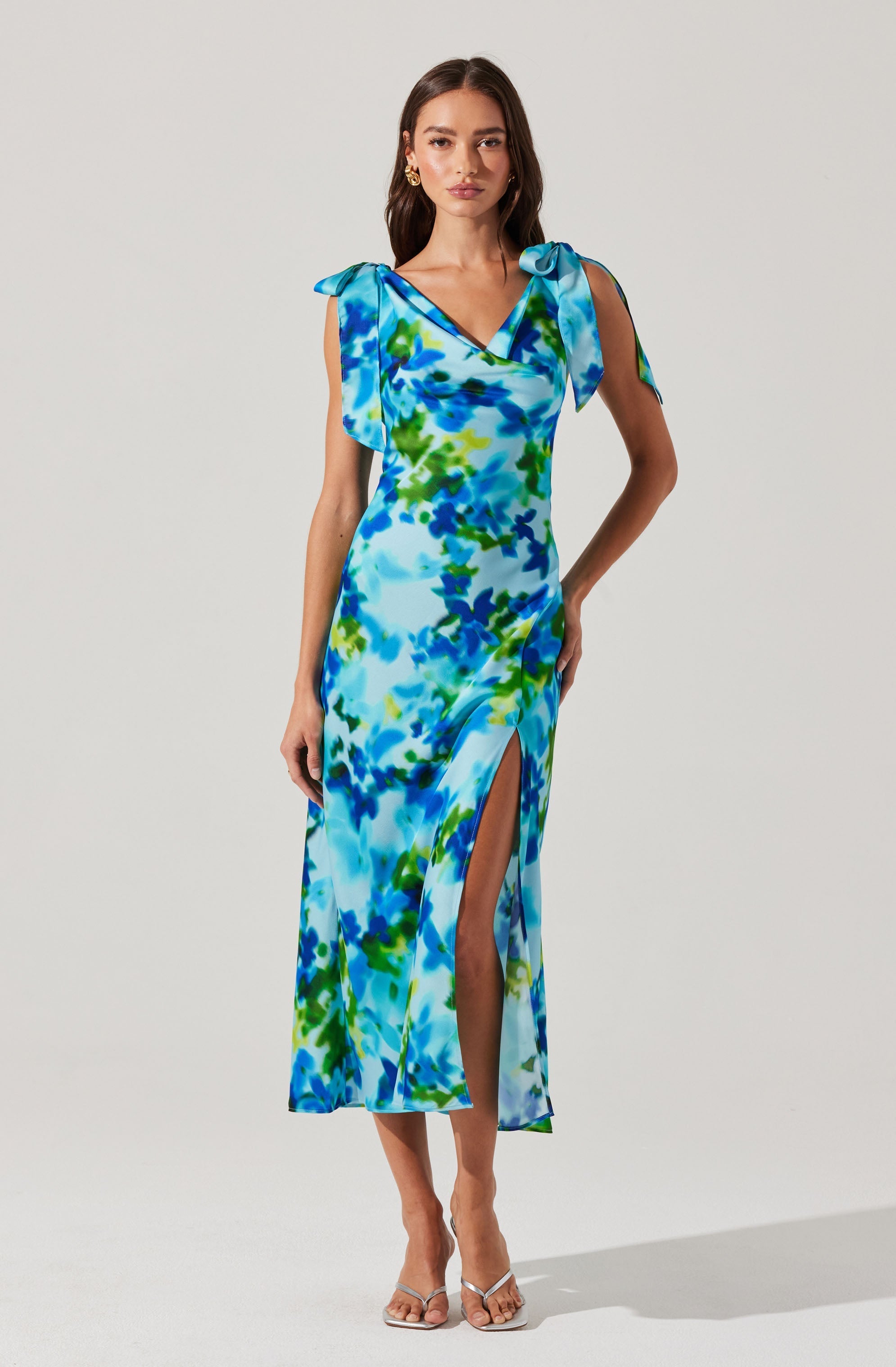 A woman is wearing a long, sleeveless dress with a vibrant, abstract watercolor pattern in shades of blue, green, and white. The dress features shoulder ties that form bows, a V-neckline, and a high slit on one side. She is also wearing clear high-heeled sandals and gold earrings.