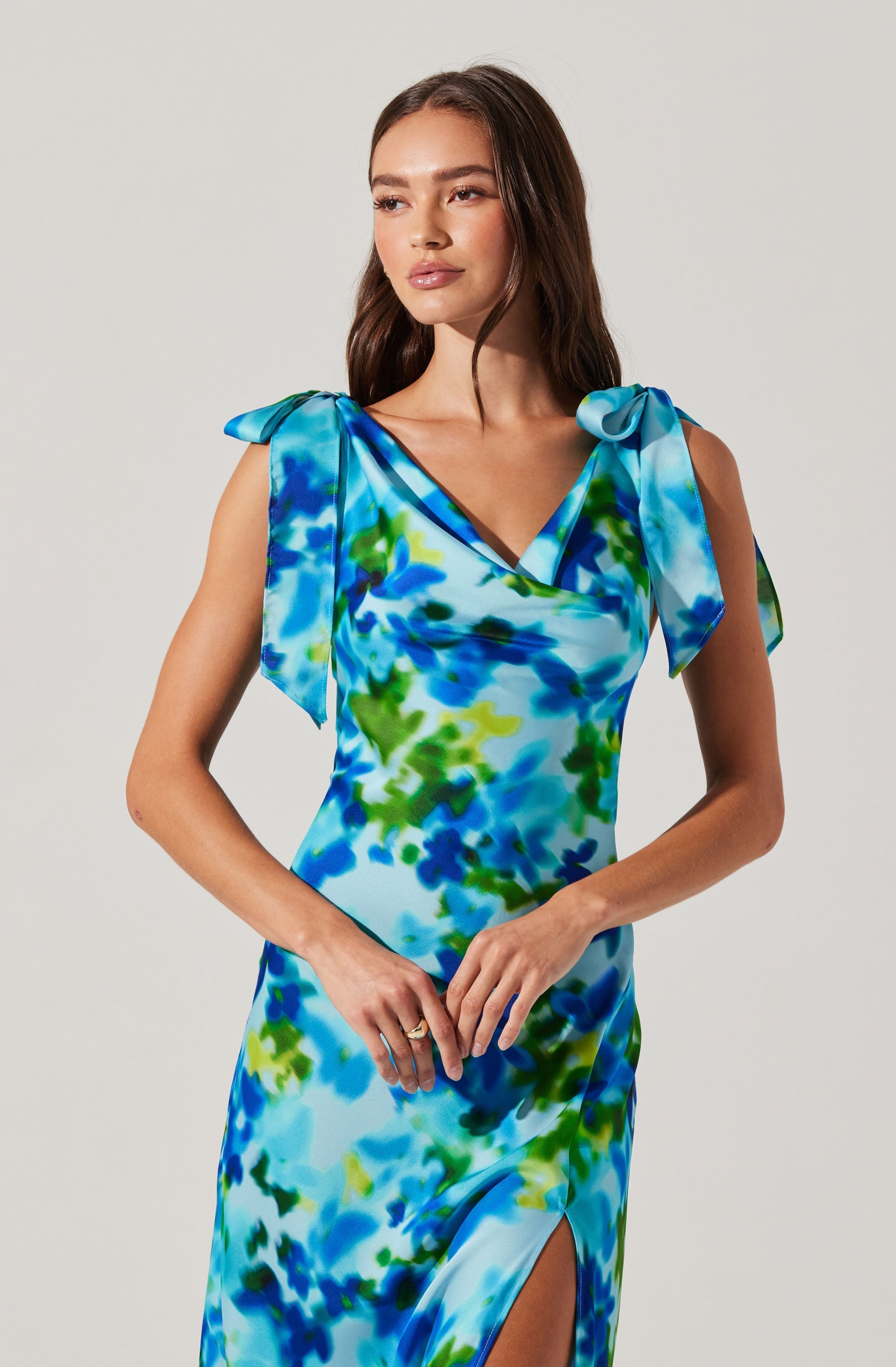 A woman is wearing a sleeveless, fitted dress with a colorful, abstract print in shades of blue, green, and white. The dress features bow-like shoulder straps and a front slit at the hem.