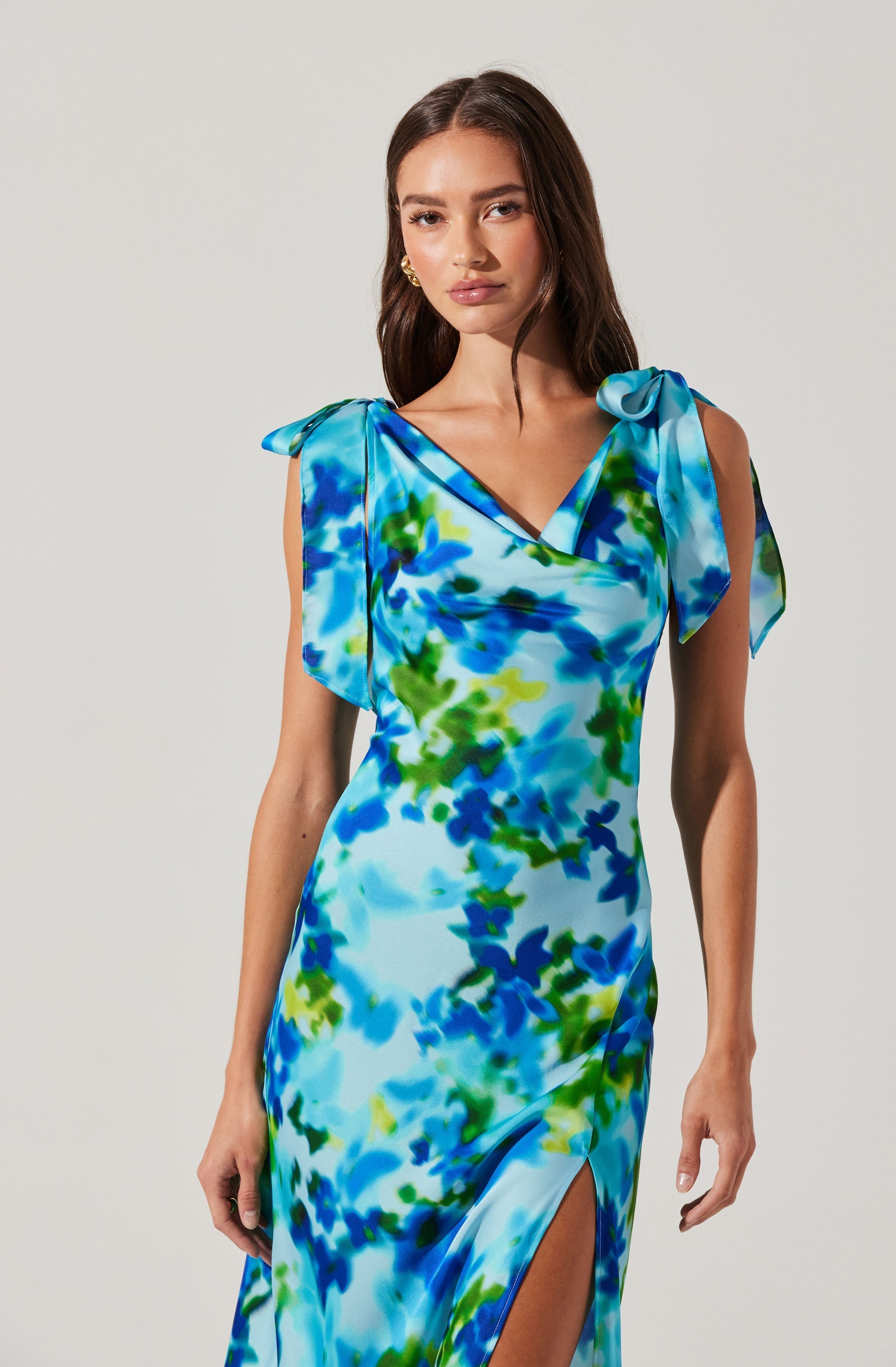 A woman is wearing a sleeveless, floor-length dress with a vibrant, abstract floral pattern in shades of blue, green, and white. The dress features draped fabric at the shoulders with tied bows on each shoulder. It has a V-neckline and a thigh-high slit on one side.