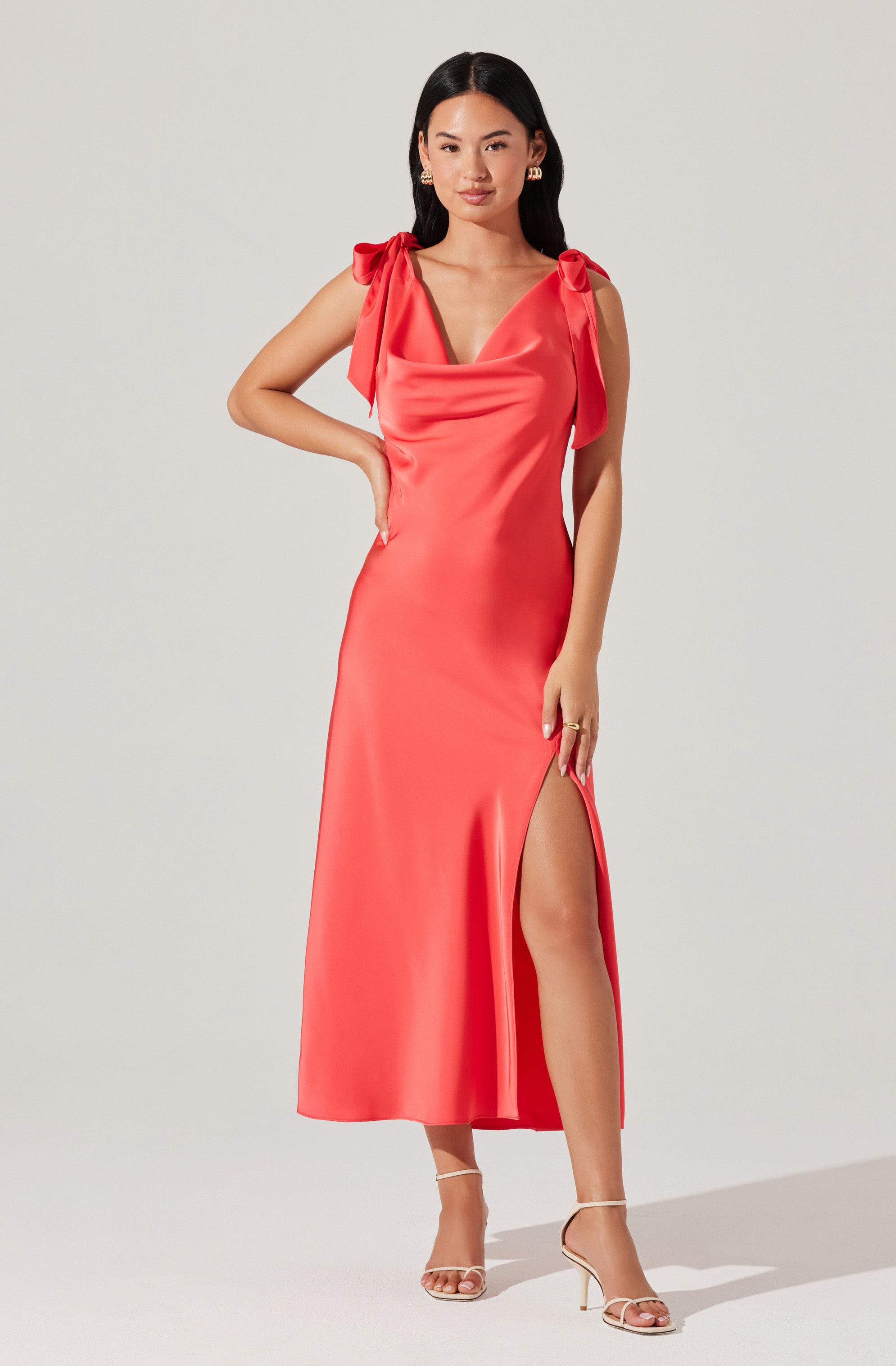 A woman is wearing a sleeveless, coral-colored satin dress with shoulder bows. The dress features a V-neckline and a high slit on one side, revealing her leg. She is also wearing nude, open-toe high heel sandals.