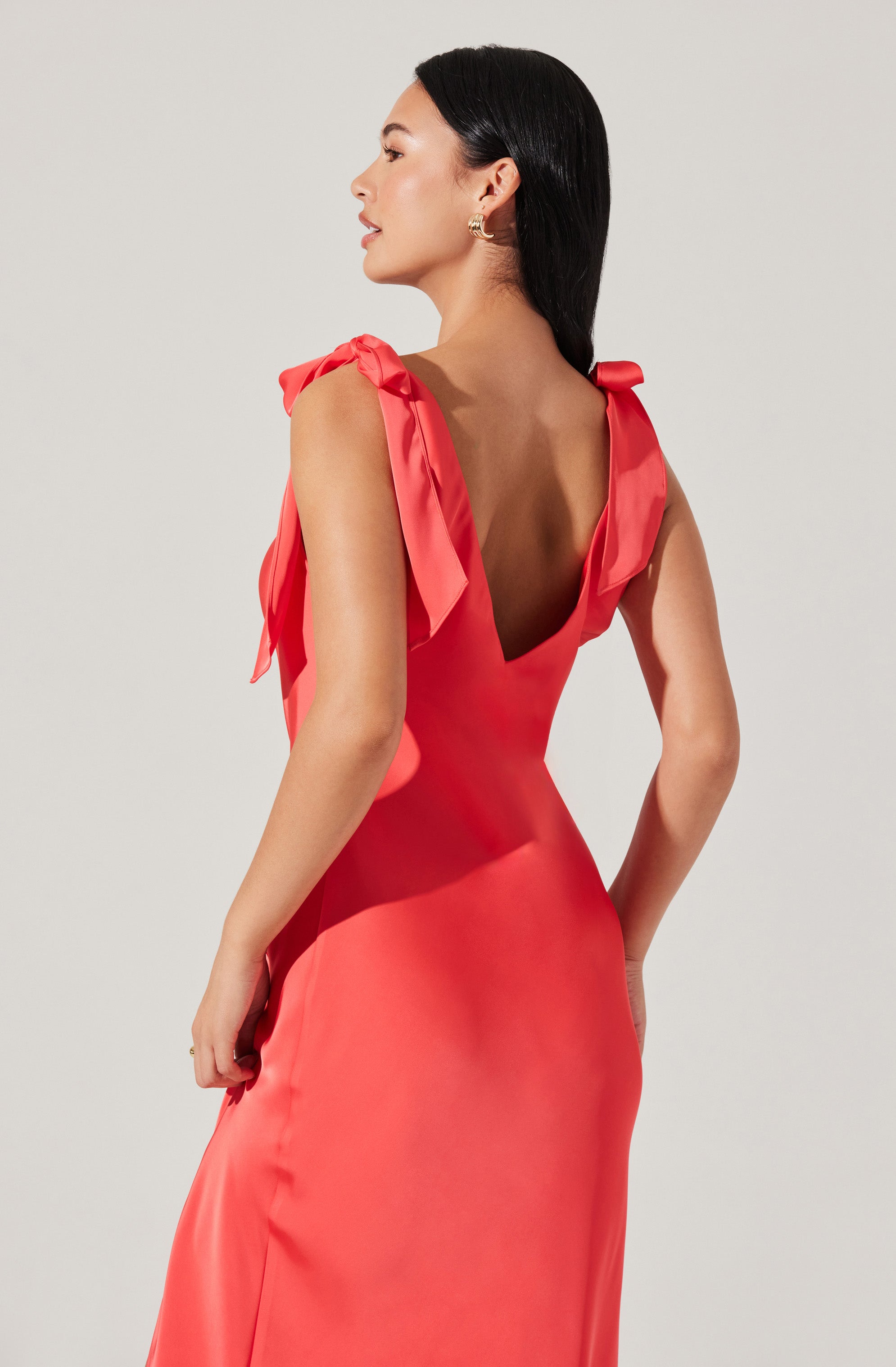 A woman is wearing a coral-colored satin dress with wide straps tied into bows on her shoulders. The dress has a deep V-neckline in the back and a fitted silhouette.