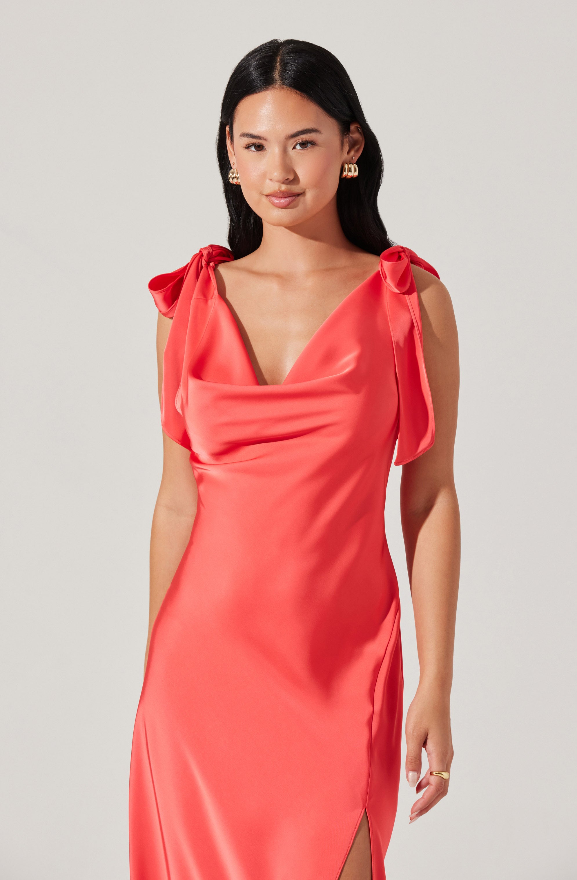 A woman is wearing a sleeveless, coral-colored satin dress with bow details on the shoulders, a deep V neckline, and a side slit at the hem.