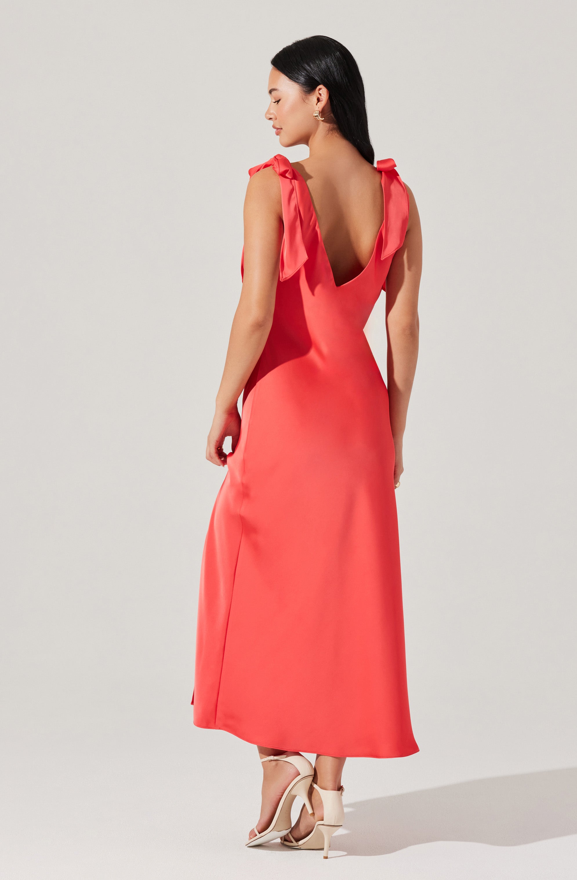 A woman is wearing a sleeveless coral-colored dress with shoulder straps tied into bows. The dress has a deep V-back design and reaches down to her ankles. She is also wearing high-heeled shoes with ankle straps.