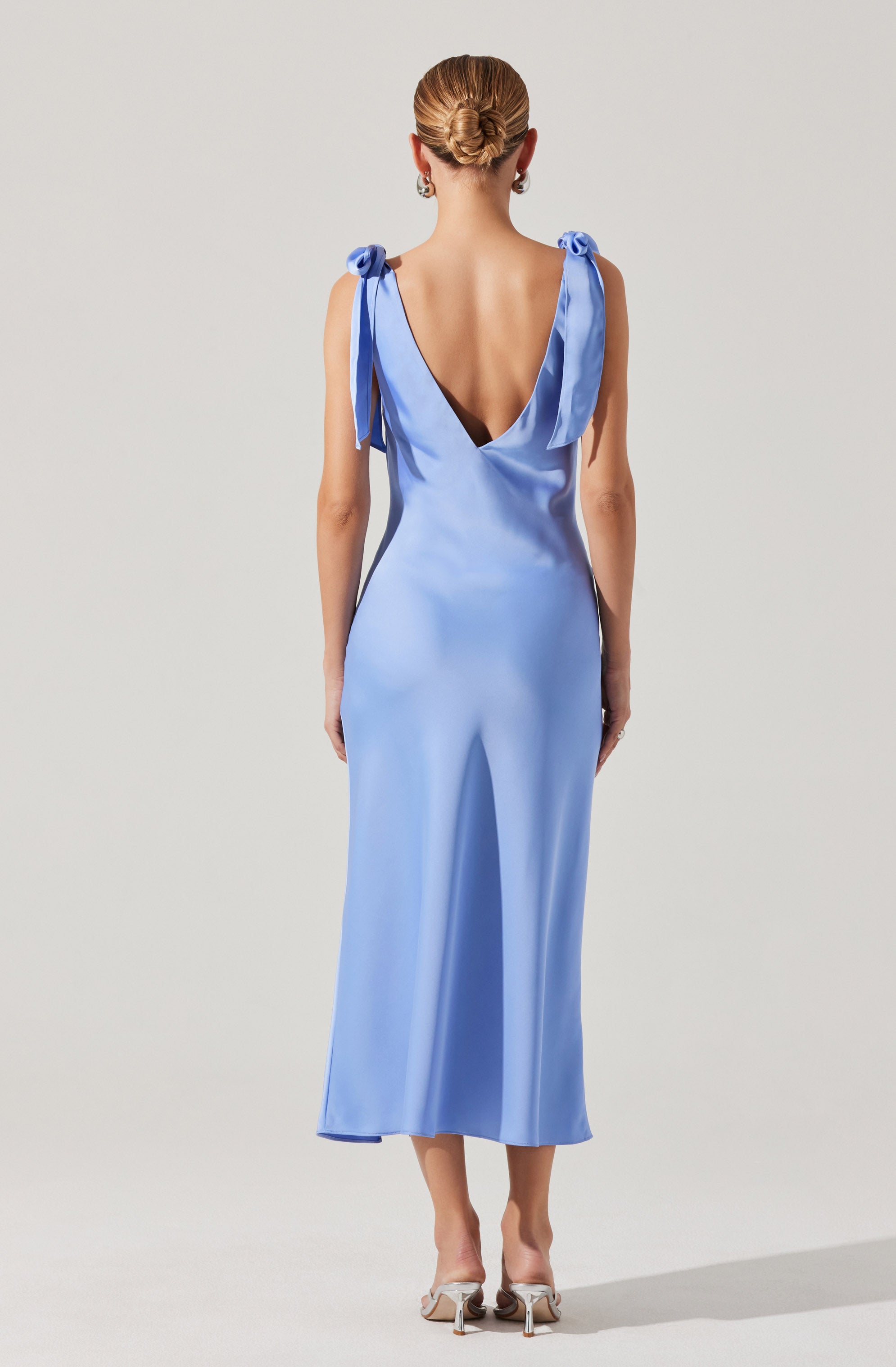 A woman is wearing a long, light blue satin dress with a deep V back. The dress features shoulder straps tied into bows. She is also wearing silver high-heeled shoes and earrings. Her hair is styled in a low, elegant bun.