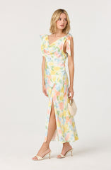 A woman is wearing a sleeveless, floor-length dress with a colorful pastel watercolor pattern. The dress has a V-neckline, ruffled shoulder details, and a front slit that reveals her leg. She is holding a small beige clutch bag and wearing nude high-heeled sandals.