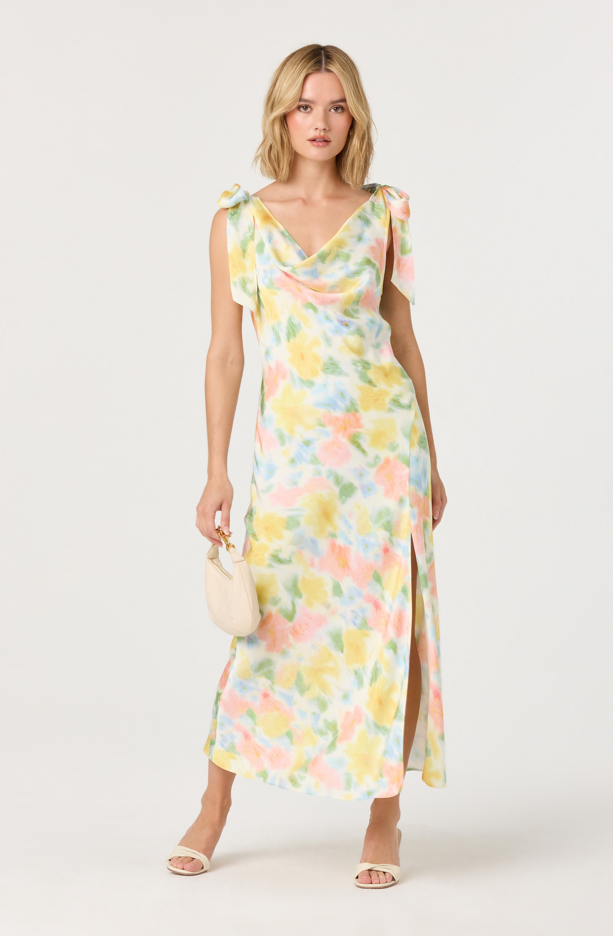 A woman is wearing a sleeveless, ankle-length dress with a colorful pastel watercolor print. The dress features a deep V neckline and shoulder ties. She is holding a small, light-colored handbag and wearing open-toe, high-heeled shoes.