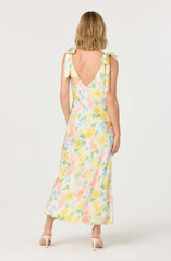 A woman is wearing a sleeveless, ankle-length dress with a watercolor floral pattern in pastel shades of yellow, pink, green, and blue. The dress features shoulder straps tied into bows on each shoulder and a deep V-shaped back. She is also wearing light-colored high-heeled shoes.