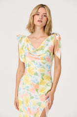 A woman is wearing a sleeveless dress with a cowl neckline and shoulder ties that resemble small bows. The dress features a colorful, watercolor-like pattern with soft pastel shades of yellow, pink, green, and blue.