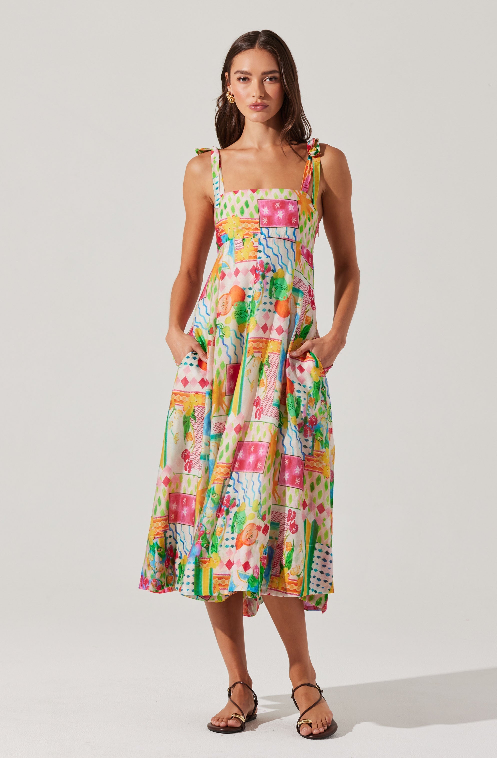 The dress is a colorful, sleeveless, midi-length piece with a square neckline, featuring adjustable shoulder straps tied into bows at the shoulders. The fabric displays a lively, multicolored pattern with various abstract and whimsical shapes, including trees, umbrellas, and other playful designs.