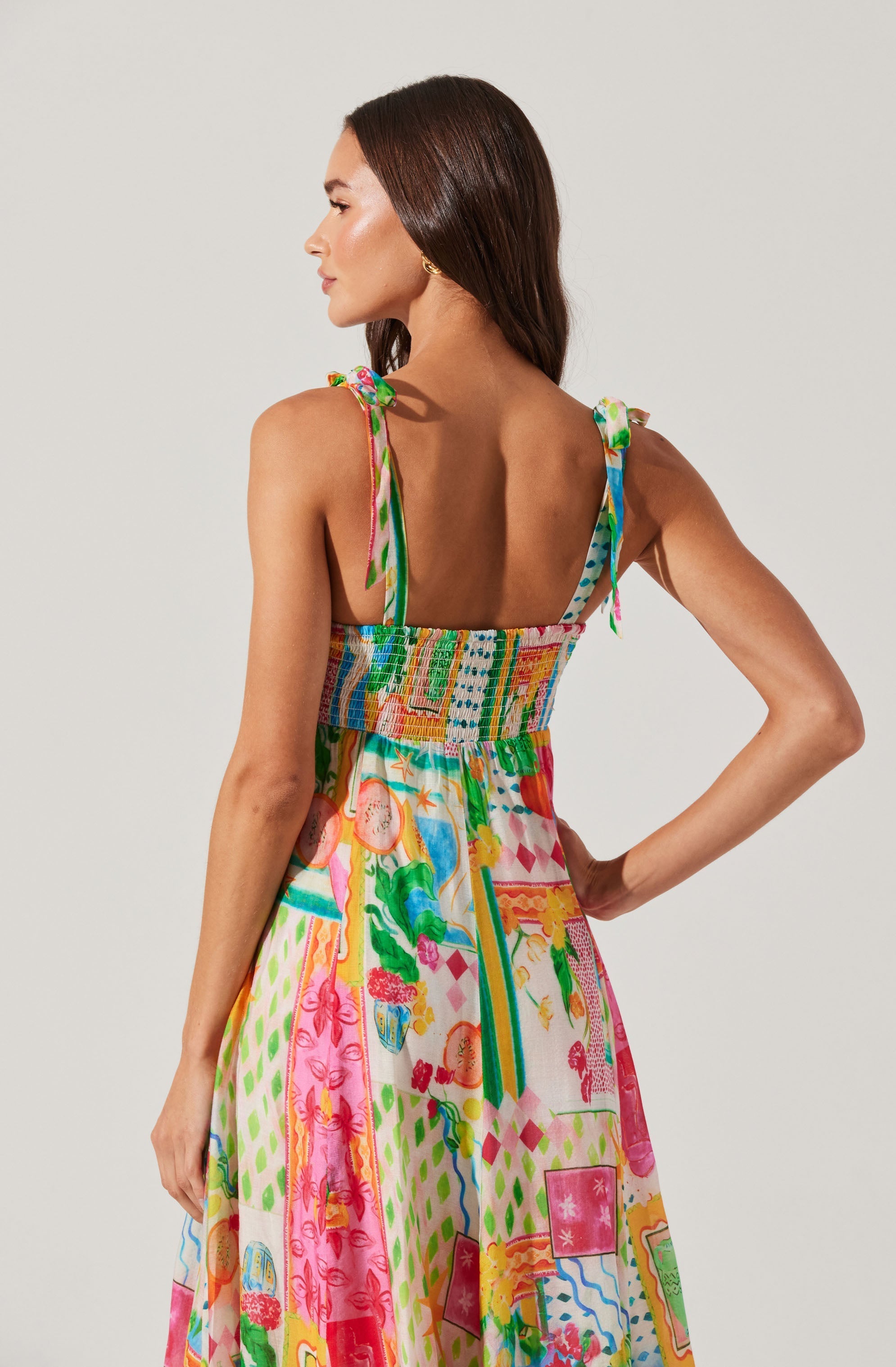 A woman is wearing a colorful, sleeveless sundress with shoulder ties. The dress features a vibrant, patchwork pattern with various abstract and floral designs in shades of pink, green, blue, yellow, and orange. The top has a smocked waist, and the fabric is lightweight and flowing.