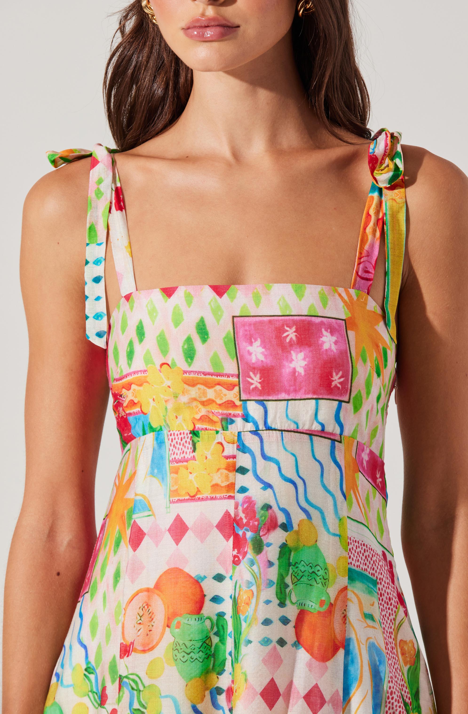 A woman is wearing a sundress with colorful, abstract patchwork patterns. The dress features wide shoulder straps with tied bows on each shoulder, and the fabric showcases various vibrant prints, including pink, green, yellow, orange, blue, and red, with geometric and playful motifs.