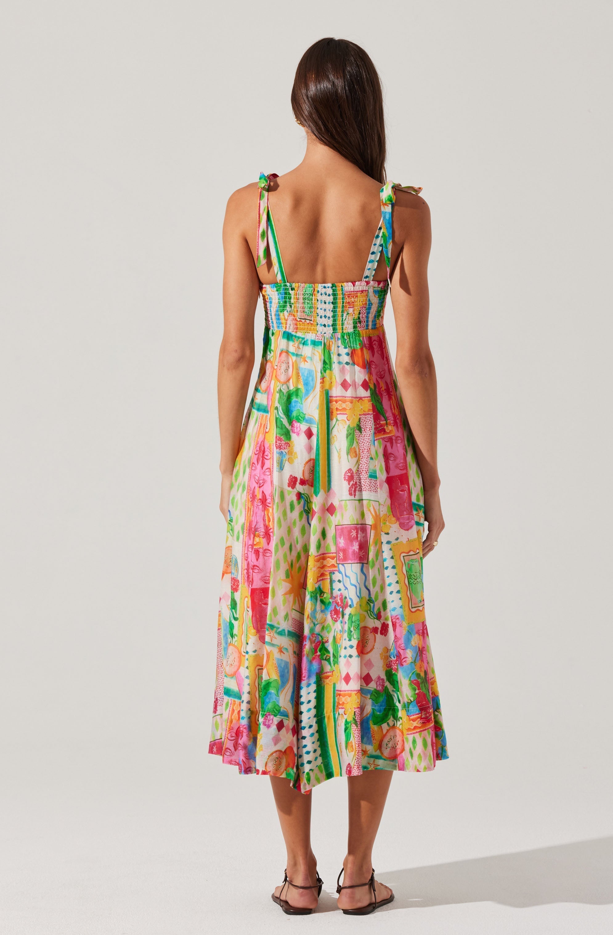 A woman is wearing a sleeveless, colorful dress with thin shoulder straps. The dress features a vibrant, abstract print with various shapes, patterns, and colors including pink, green, blue, yellow, and orange. The fabric appears lightweight and the length reaches mid-calf. The woman's hair is long, straight, and brown, and she is standing against a plain, light-colored background.