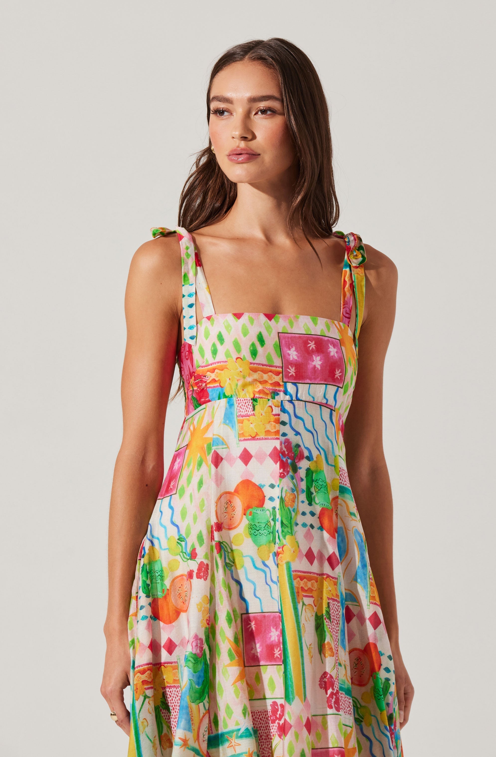 A woman is wearing a sleeveless dress with shoulder straps tied into bows. The dress features a vibrant, colorful print with various abstract shapes, patterns, and images in shades of pink, green, orange, yellow, blue, and white.