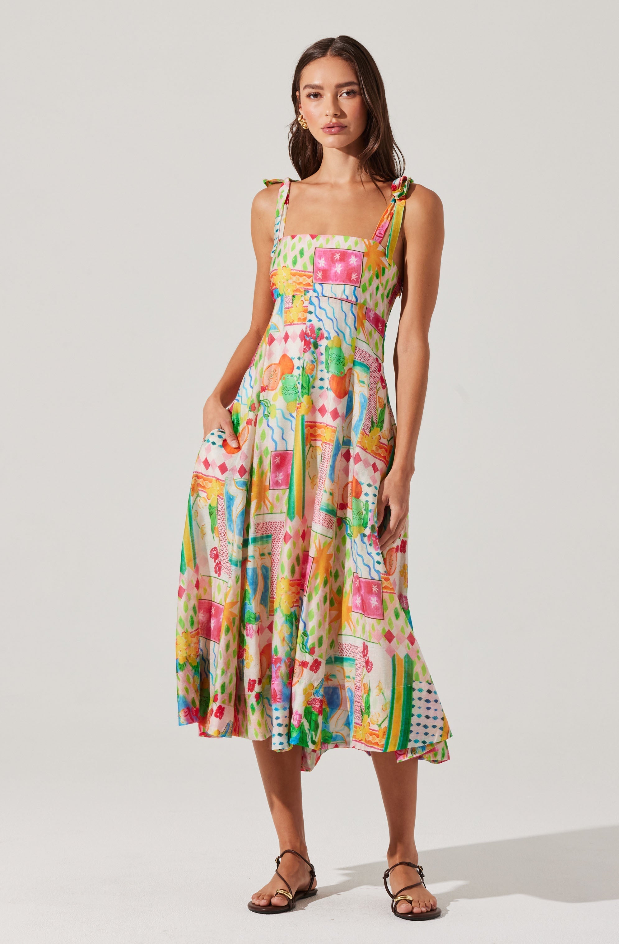 A woman is wearing a colorful, patterned sundress with a fitted bodice and a flowing, midi-length skirt. The dress has thin, adjustable shoulder straps with small bows on the shoulders. The pattern includes various geometric shapes, florals, and abstract designs in bright colors such as pink, yellow, green, blue, and orange. She is also wearing simple, strappy sandals.