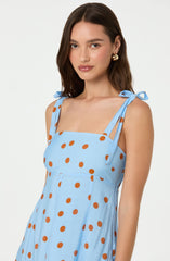 A woman is wearing a sleeveless light blue dress with brown polka dots. The dress features shoulder tie straps that form small bows.