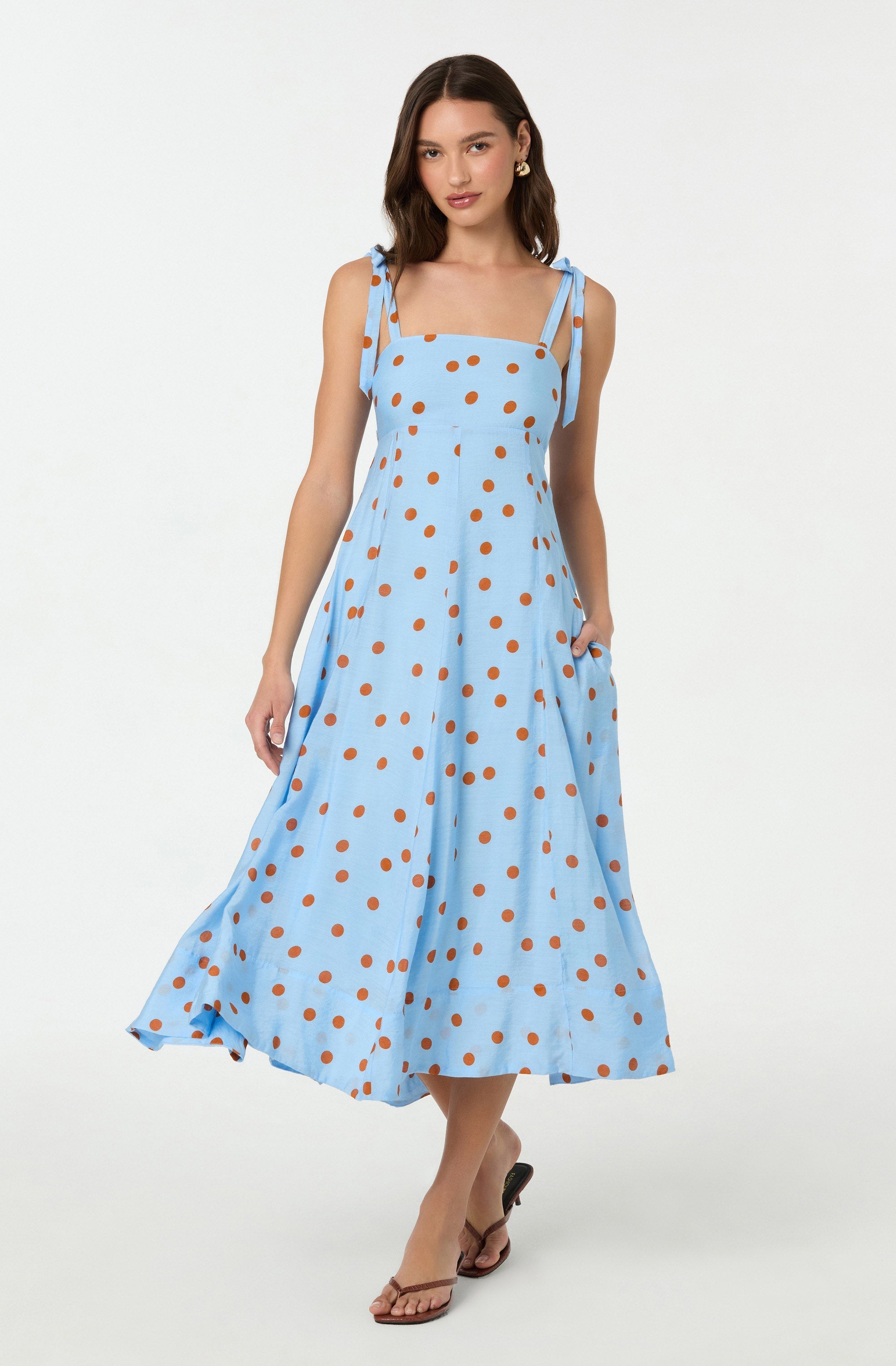 A woman is wearing a light blue sleeveless dress with thin, tied shoulder straps. The dress features a pattern of evenly spaced, large brown polka dots and has a fitted bodice with a gathered waist, flowing into a full, ankle-length skirt. She is also wearing tan open-toe sandals with a slight heel. The background is plain white.