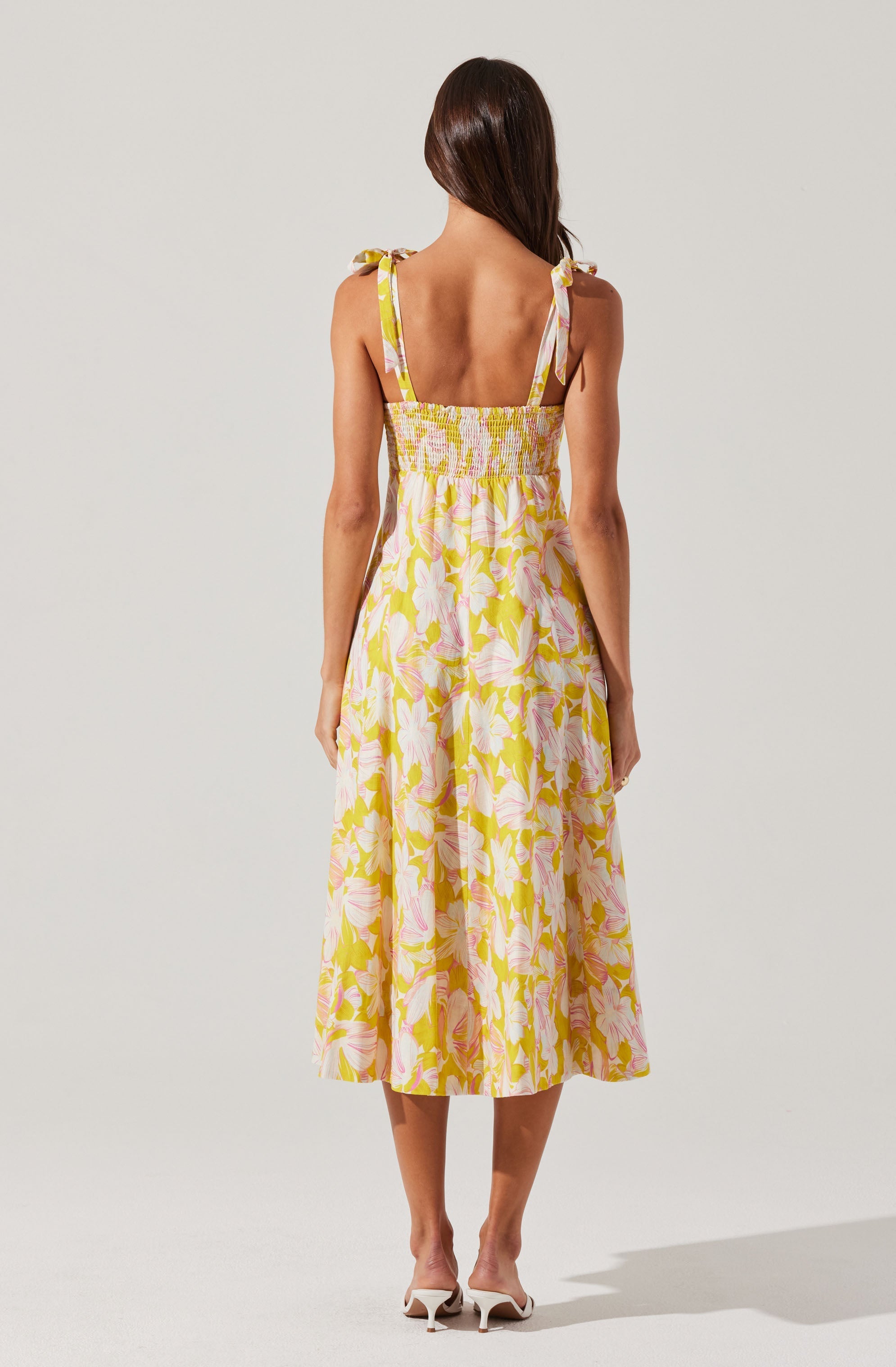 The dress features shoulder straps tied into bows at the shoulders, a smocked waistline, and a flowing, A-line skirt that falls below the knees. The fabric has a floral pattern with large pink flowers and green leaves on a light background.