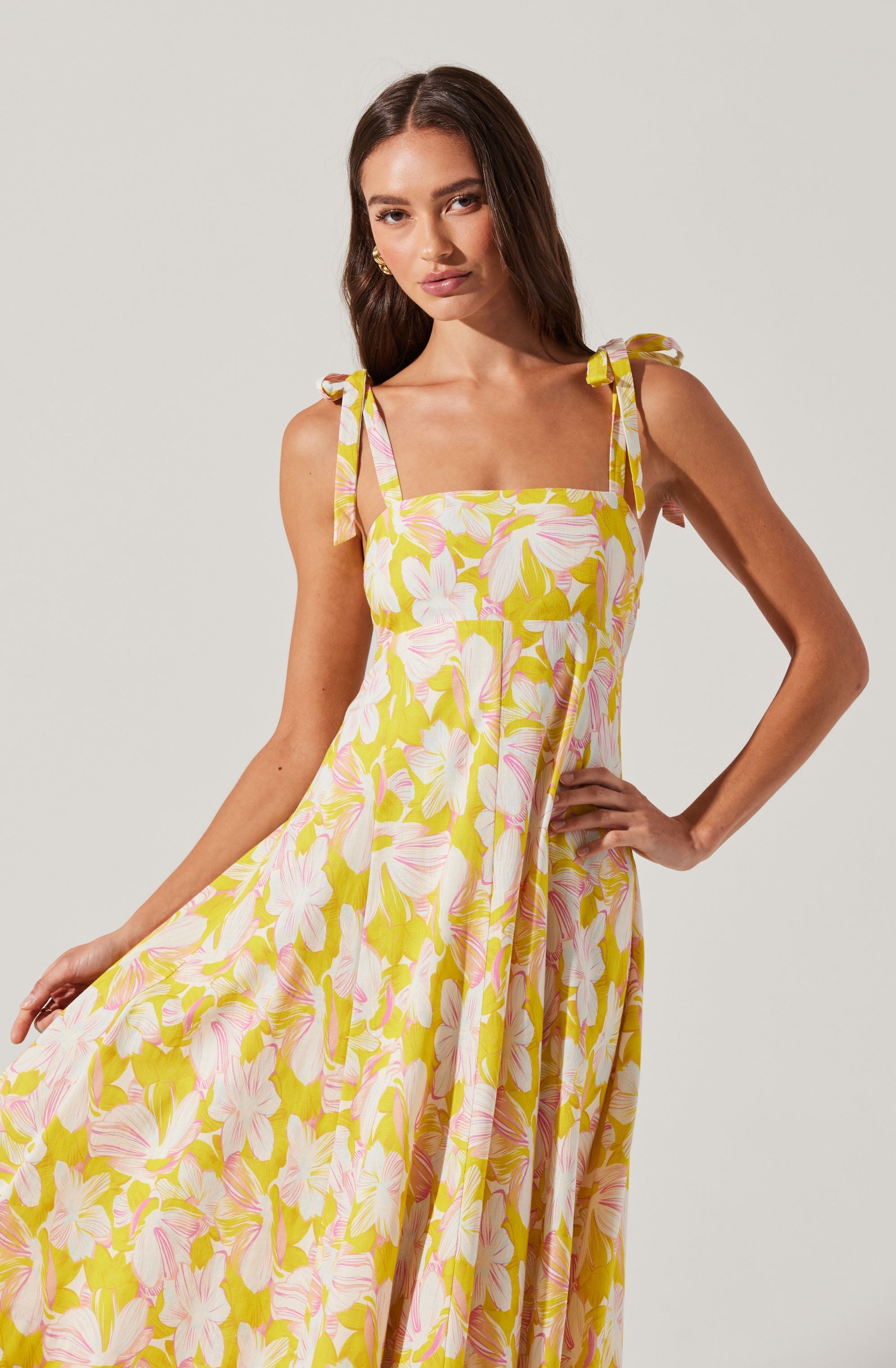 A woman is wearing a sleeveless, floor-length dress with a floral pattern. The dress features pink and white flowers with green leaves on a yellow background. It has thin shoulder straps tied into bows at the shoulders, and a square neckline. The dress has a relaxed, flowing fit.