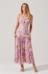 A woman is wearing a strapless, floral maxi dress with a fitted bodice that has a twisted knot detail at the top and a small front slit. The dress features a pink, purple, and green floral pattern on a light, sheer fabric. The bottom part is ruffled and semi-transparent, reaching her ankles. She is also wearing nude high-heeled sandals.