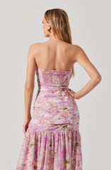 A woman is wearing a strapless, floor-length dress with a pink floral pattern. The upper part of the dress features smocking detail, and the fabric has a soft, flowing appearance with gathered sections near the hips and a ruffled hemline at the bottom. The dress is predominantly pink with hints of green and white in the floral design.