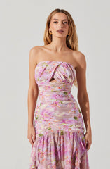 A woman is wearing a strapless, floral dress with a pink, purple, and green pattern. The dress features a cut-out detail at the bust, pleated fabric, and a gathered section at the bottom with a ruffled hem. She is also wearing gold earrings.