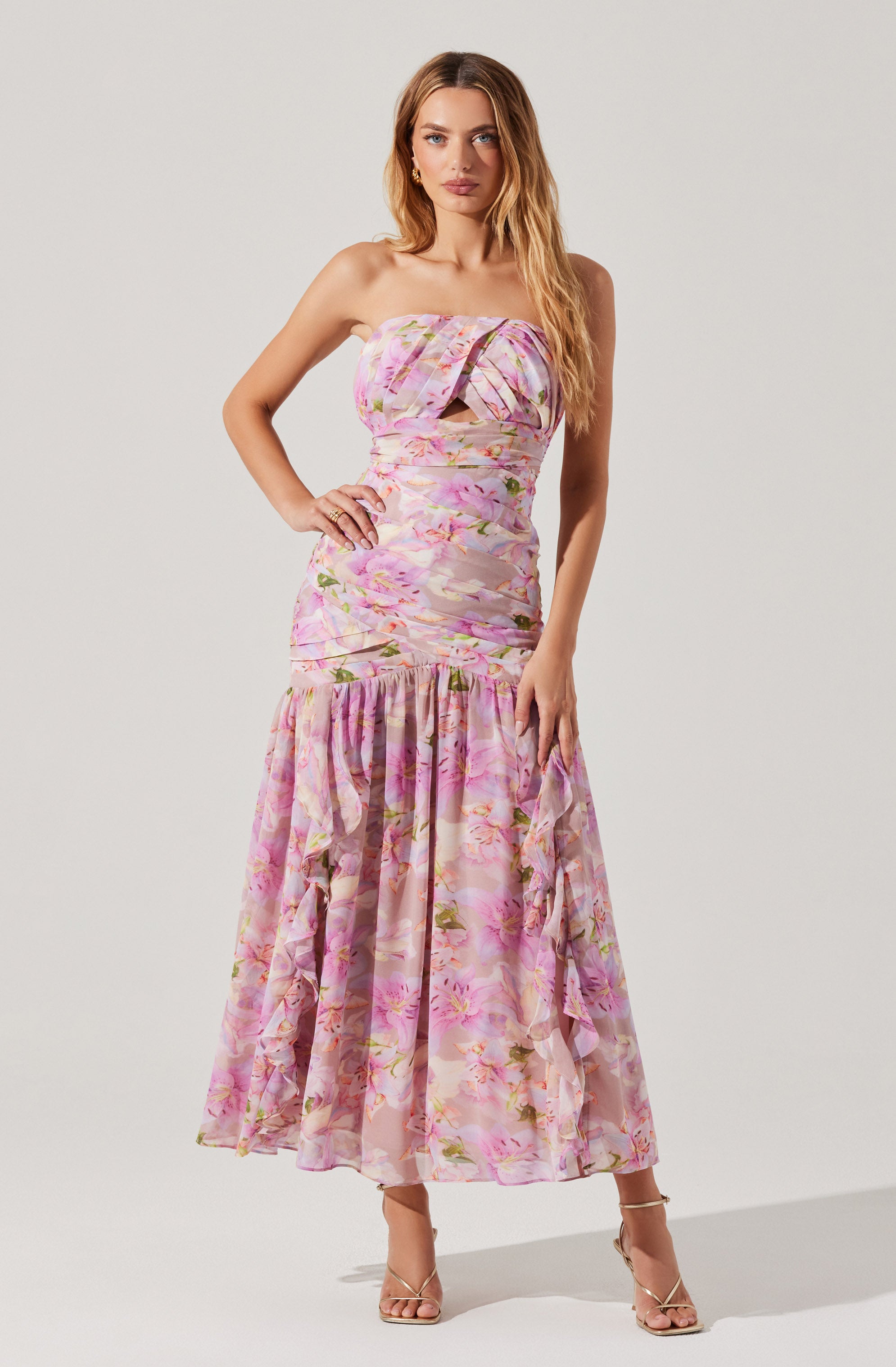 A woman is wearing a strapless, floor-length dress with a pink, purple, and green floral pattern. The dress features a ruched bodice with a large bow detail at the center and a ruffled, flowing skirt. She is also wearing open-toe high heel sandals.