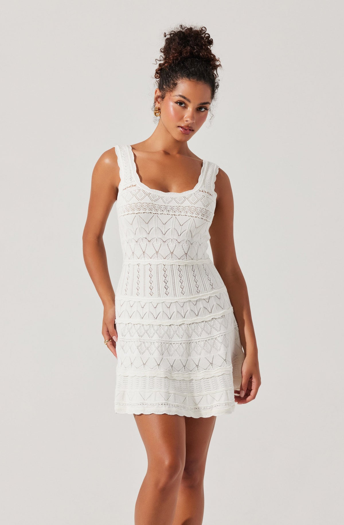 A woman is wearing a sleeveless white dress with a square neckline. The dress features a textured, embroidered pattern with various geometric designs, including diamonds and zigzags, arranged in horizontal sections. The fabric has a lace-like appearance, giving the dress a delicate and airy look. The dress falls above the knee and has a fitted waist.