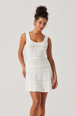 A woman is wearing a sleeveless white dress with a square neckline. The dress features a textured, embroidered pattern with various geometric designs, including diamonds and zigzags, arranged in horizontal sections. The fabric has a lace-like appearance, giving the dress a delicate and airy look. The dress falls above the knee and has a fitted waist.