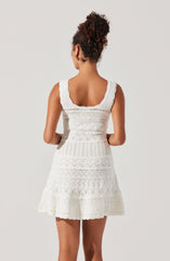 A woman is wearing a sleeveless, white dress with a fitted waist and a flared skirt. The dress features intricate lace and embroidered patterns throughout, with a scalloped hemline and wide, scalloped shoulder straps.