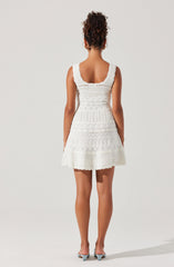 A woman is wearing a sleeveless, knee-length white dress with lace and embroidered patterns. The dress has wide straps and a scoop neckline at the back. She is also wearing light-colored high-heeled shoes.