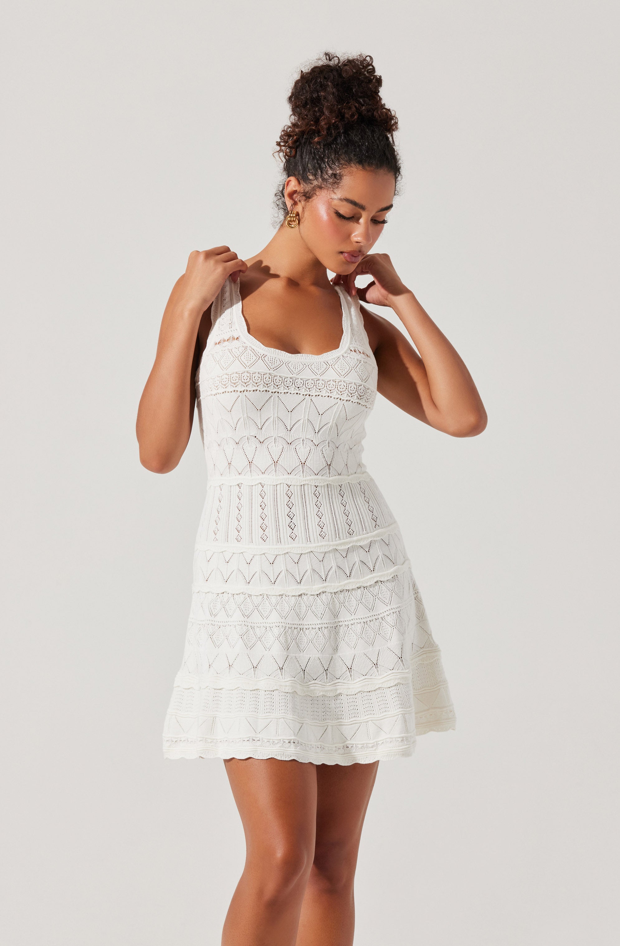 A woman is wearing a sleeveless, knee-length white dress with intricate lace and embroidery patterns. The dress has a fitted bodice with a scoop neckline and layered sections with different lace designs, creating a textured appearance. She has her hair styled in a curly updo and is holding the straps of her dress with both hands, looking down with a contemplative expression.