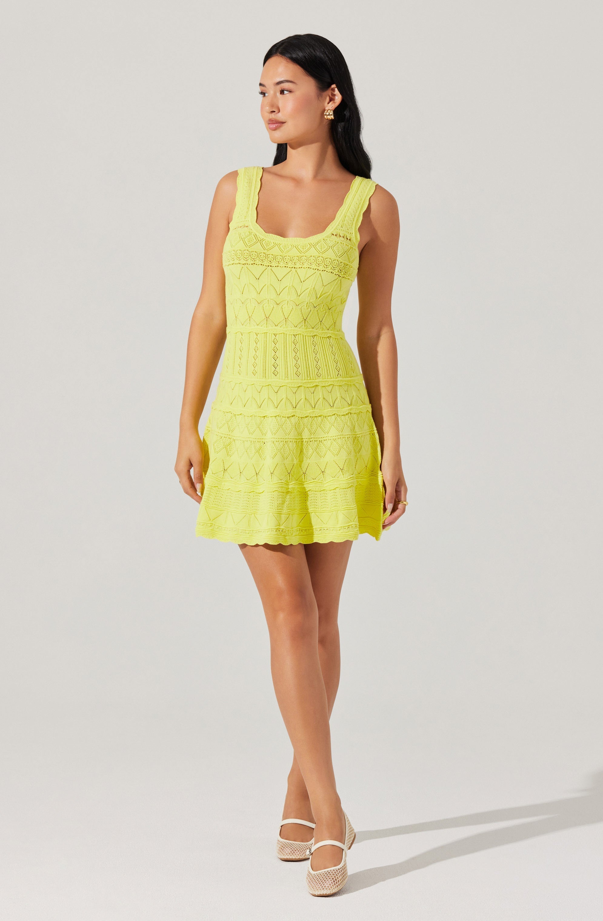 A woman is wearing a sleeveless, above-the-knee yellow dress with intricate lace and embroidered patterns throughout. The dress has wide shoulder straps and a fitted waist. She is also wearing white high-heeled shoes with ankle straps.