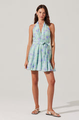 A woman is wearing a sleeveless, halter-neck dress with a pastel watercolor pattern in shades of light blue, lavender, and green. The dress has a relaxed fit with a waistband tied with a bow at the waist and a gathered, flowing skirt that ends above the knees. She is paired with white, flat sandals with ankle straps.