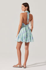 A woman is wearing a sleeveless dress with a halter neckline tied at the back with a bow. The dress features a light, watercolor-like pattern in shades of green, purple, and blue. The skirt of the dress is flared and above the knee length. She is also wearing white sandals with ankle straps.