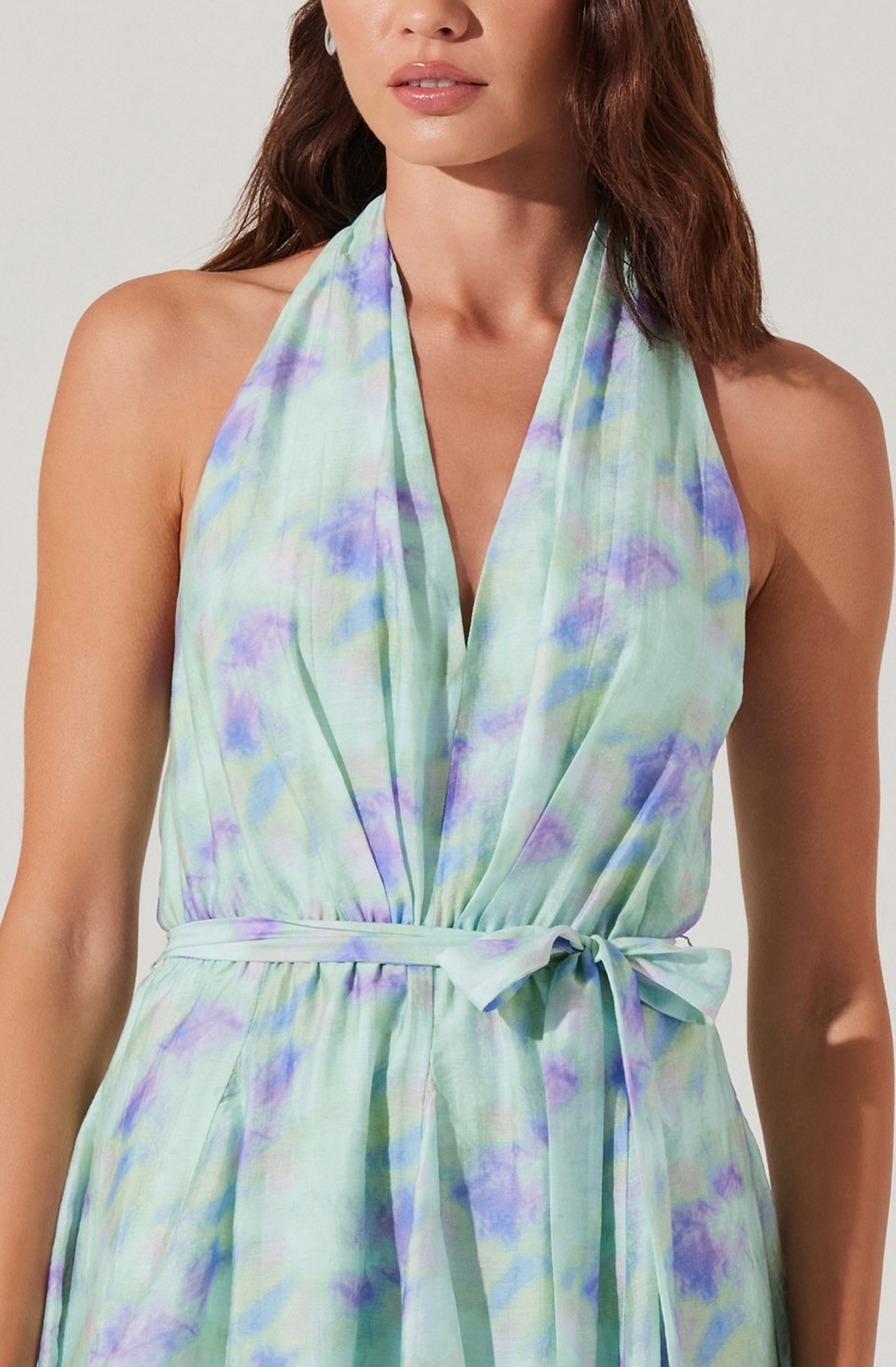 A woman is wearing a sleeveless, halter-style dress with a soft watercolor pattern in shades of light green, purple, and blue. The dress features a waist tie bow, matching the fabric, that cinches at the waist. The fabric appears lightweight and flowy, suitable for warm weather.