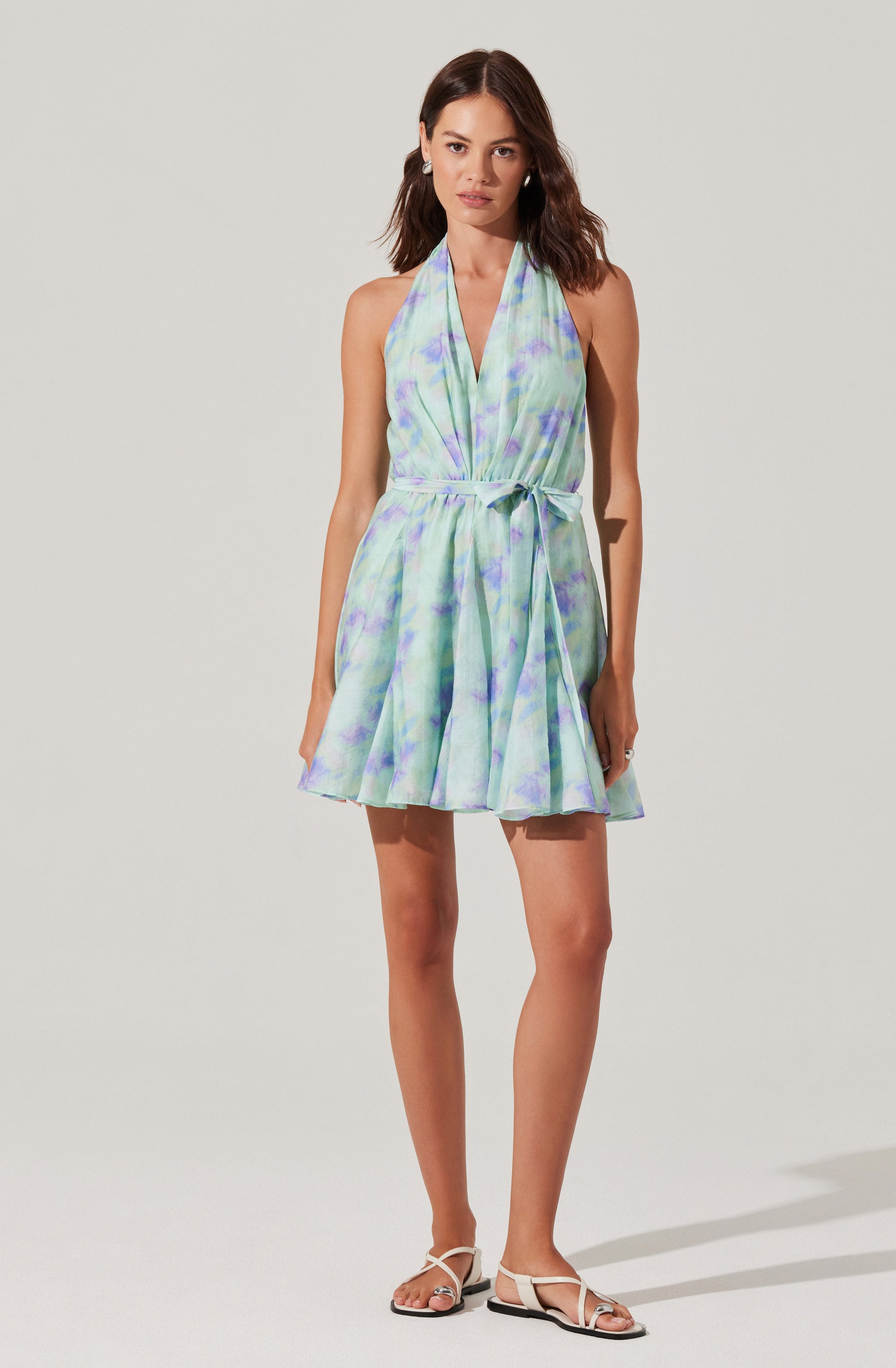 A woman is wearing a sleeveless, halter-neck dress with a pastel tie-dye pattern in shades of green, blue, and purple. The dress has a fitted waist with a fabric belt tied in a bow on the side and a flared, knee-length skirt. She is also wearing white strappy sandals.