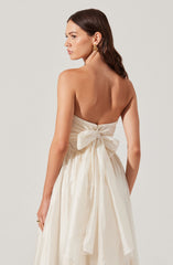 A woman is wearing a strapless, off-white, satin dress with a large bow tied at the back. The dress has a gathered bodice and a flowing skirt. She has shoulder-length dark hair, gold earrings, and a ring on her left hand.