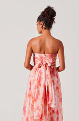 A woman is wearing a strapless, flowing dress with a watercolor-like pattern in shades of pink, orange, and white. The dress features a large bow at the back, tied at the waist, with fabric draping down. She has her hair styled in a high, curly bun and is accessorized with gold hoop earrings.