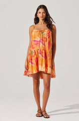 The dress is a sleeveless, above-the-knee style with thin adjustable spaghetti straps. It features a vibrant, multicolored abstract pattern predominantly in shades of orange, pink, and green. The dress has a loose, flowy fit with a ruffled hemline, creating a casual and summery look.