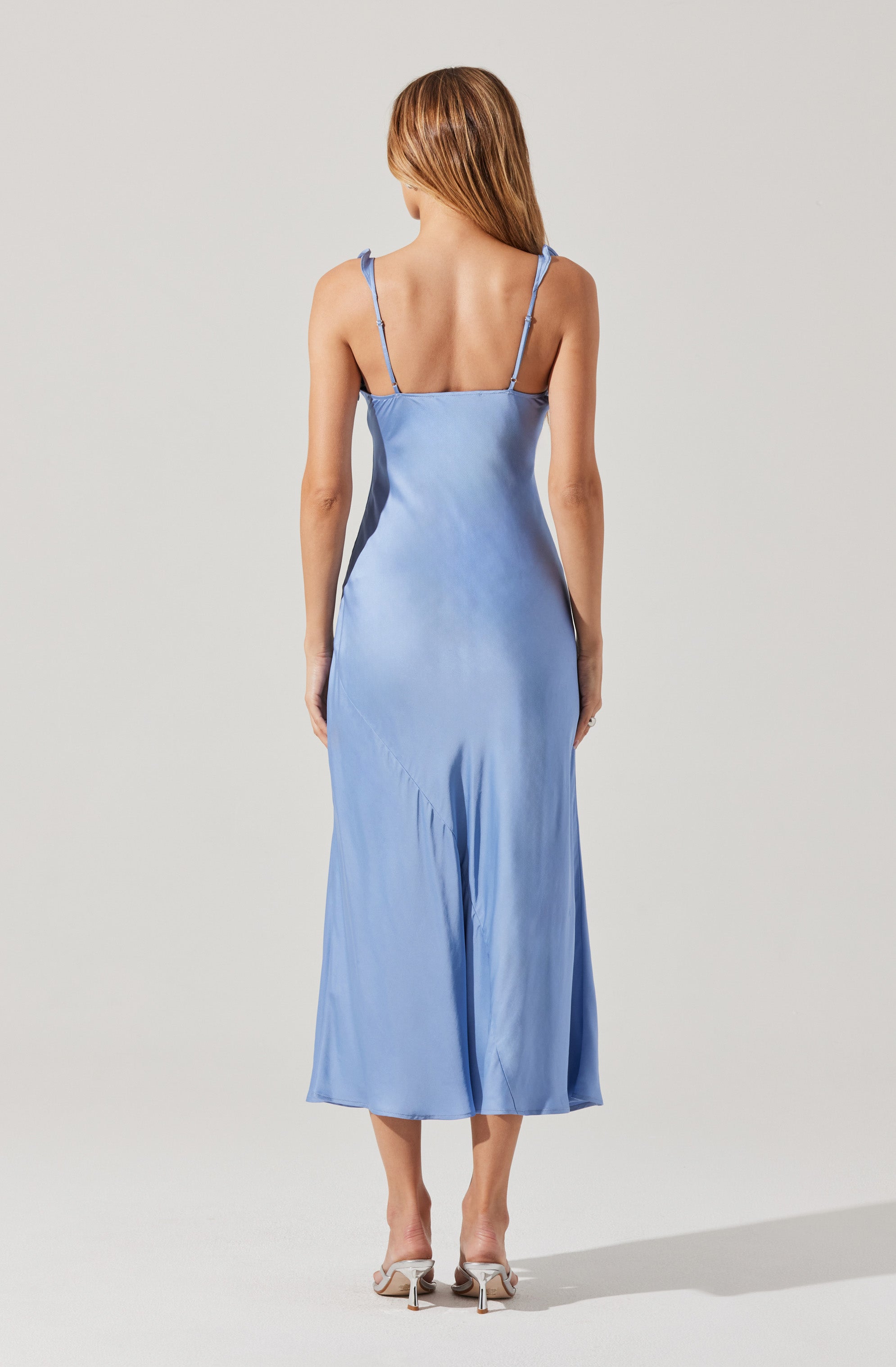 A woman is wearing a light blue satin slip dress with spaghetti straps and a fitted bodice, flowing down to a midi length. She pairs the dress with beige high-heeled shoes.