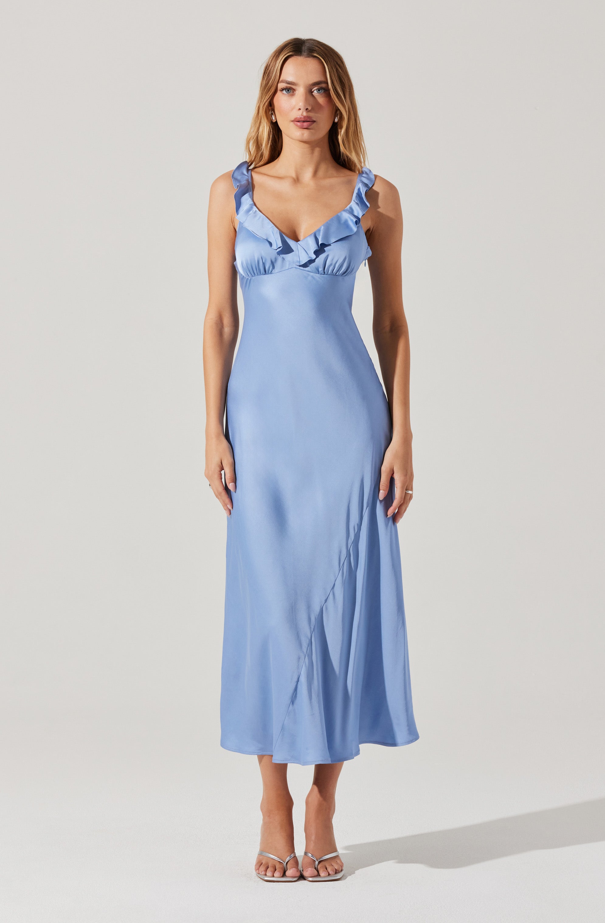A woman is wearing a long, light blue satin dress with ruffled straps and a gathered detail at the bust. The dress fits snugly at the waist and flows down to the floor with a slight sheen and a subtle slit. She is also wearing open-toe, strappy high-heeled sandals.