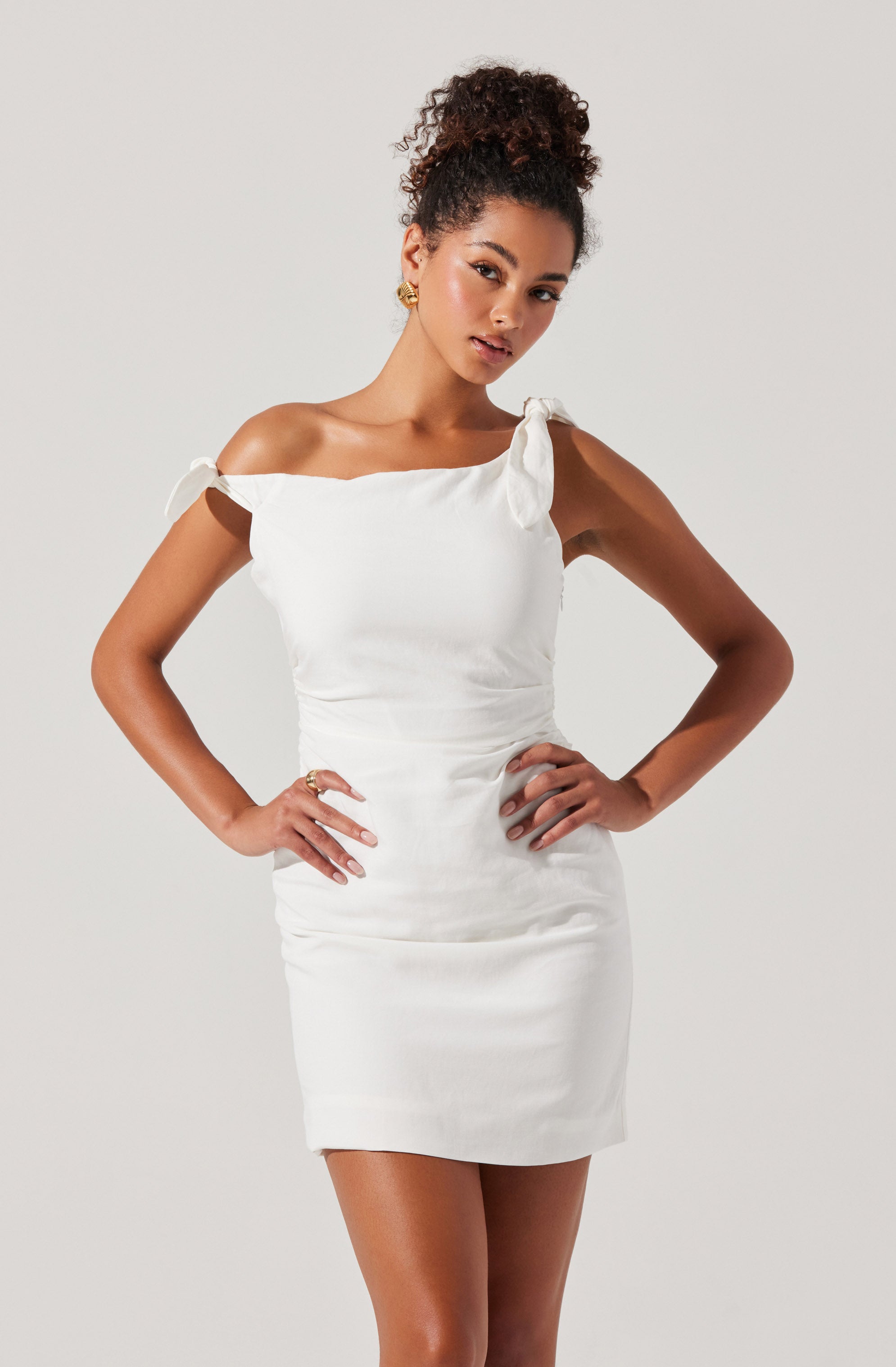 A woman is wearing a white, form-fitting dress that features off-the-shoulder sleeves tied into small bows on each shoulder. The dress is smooth and has a slightly gathered design at the waist.