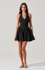 A woman is wearing a sleeveless black dress with a plunging V-neckline and a fitted bodice. The skirt is full and flared with gathered fabric, creating a voluminous and playful look. She is also wearing a pair of strappy high-heeled sandals.