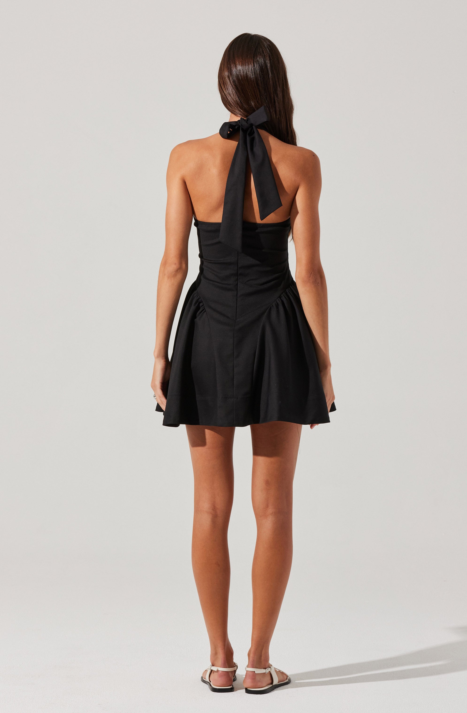 A woman is wearing a black strapless dress with a fitted bodice and a flared skirt that has gathered details at the sides. The dress features a large black ribbon tied into a bow at the back of the neck, and she is wearing flat, open-toed sandals with ankle straps.