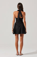A woman is wearing a black strapless dress with a fitted bodice and a flared skirt that has gathered details at the sides. The dress features a large black ribbon tied into a bow at the back of the neck, and she is wearing flat, open-toed sandals with ankle straps.