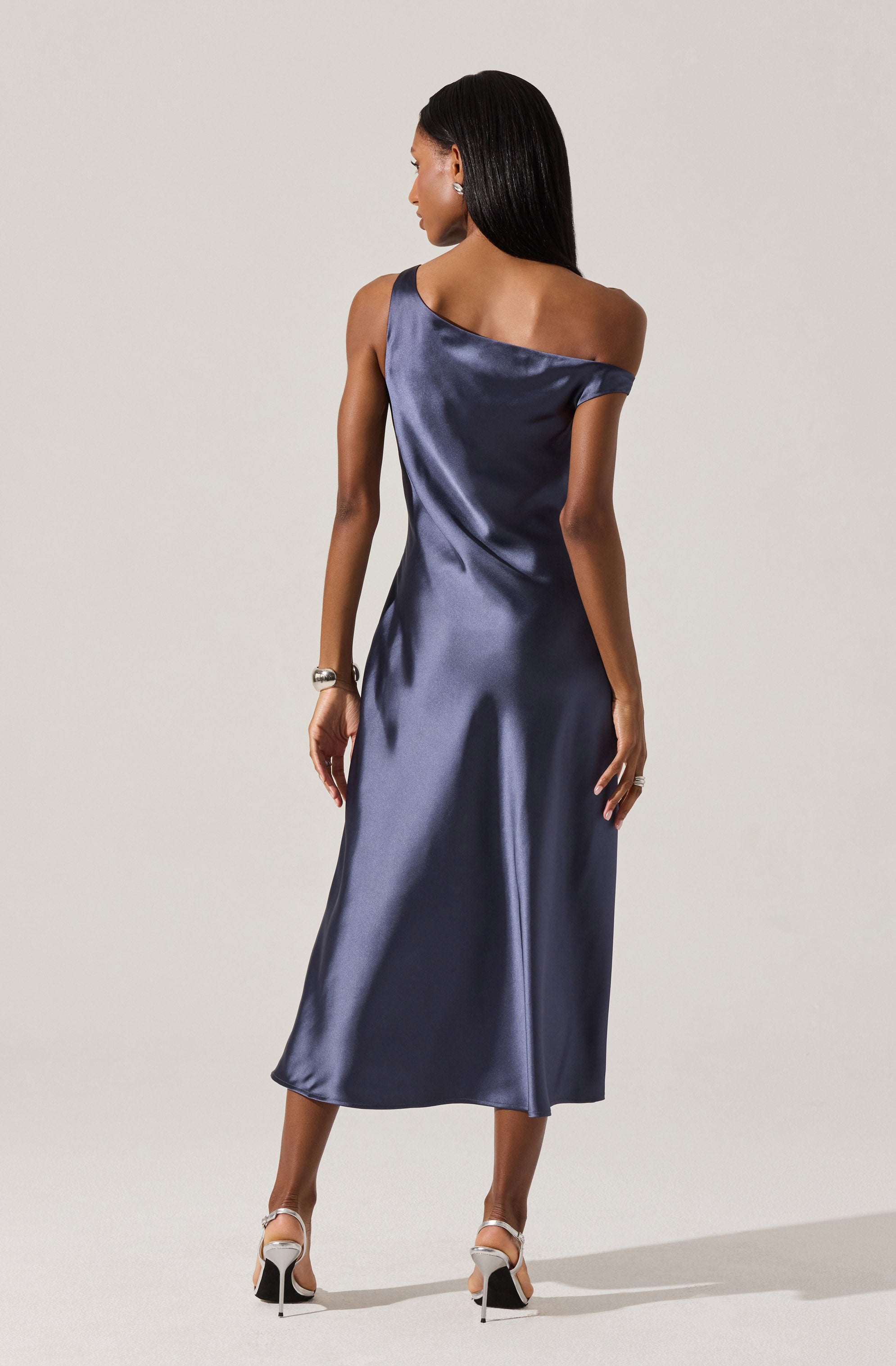 A woman is wearing a floor-length, satin dress in a deep navy blue color. The dress features a one-shoulder design with a draped, off-the-shoulder sleeve on one side and a sleeveless strap on the other. The fabric has a shiny, smooth appearance with a slight sheen, and it falls elegantly to her ankles. She is accessorized with a bracelet on her left wrist and a ring on her right hand, and she is wearing high-heeled shoes with ankle straps.