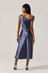 A woman is wearing a floor-length, satin dress in a deep navy blue color. The dress features a one-shoulder design with a draped, off-the-shoulder sleeve on one side and a sleeveless strap on the other. The fabric has a shiny, smooth appearance with a slight sheen, and it falls elegantly to her ankles. She is accessorized with a bracelet on her left wrist and a ring on her right hand, and she is wearing high-heeled shoes with ankle straps.