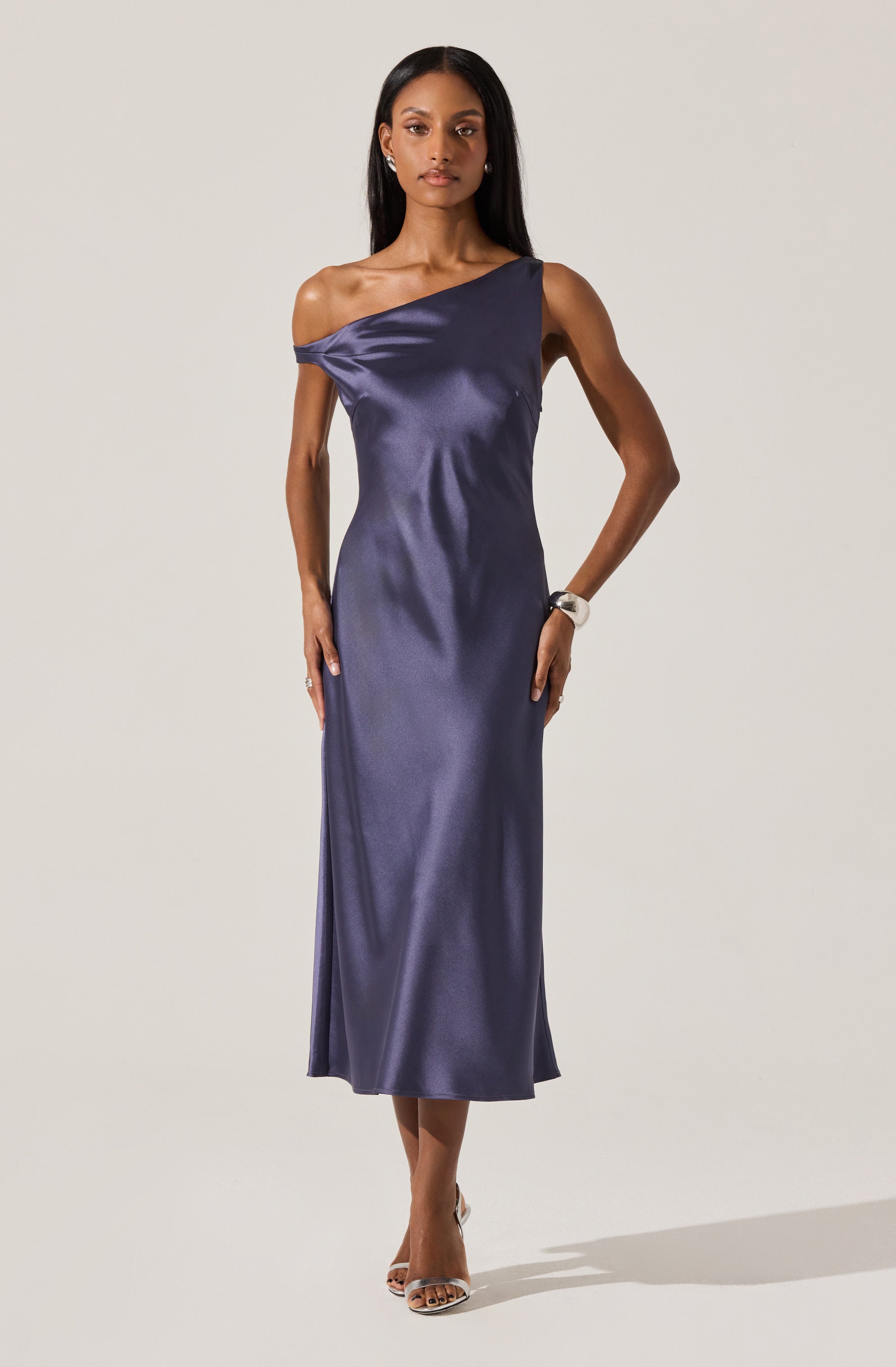 A woman is wearing a dark blue, satin, one-shoulder gown that drapes over her body, reaching down to her ankles. The dress features a loose, elegant fit with a single off-the-shoulder strap on the left side. She is accessorized with earrings, a watch, and open-toe heels.