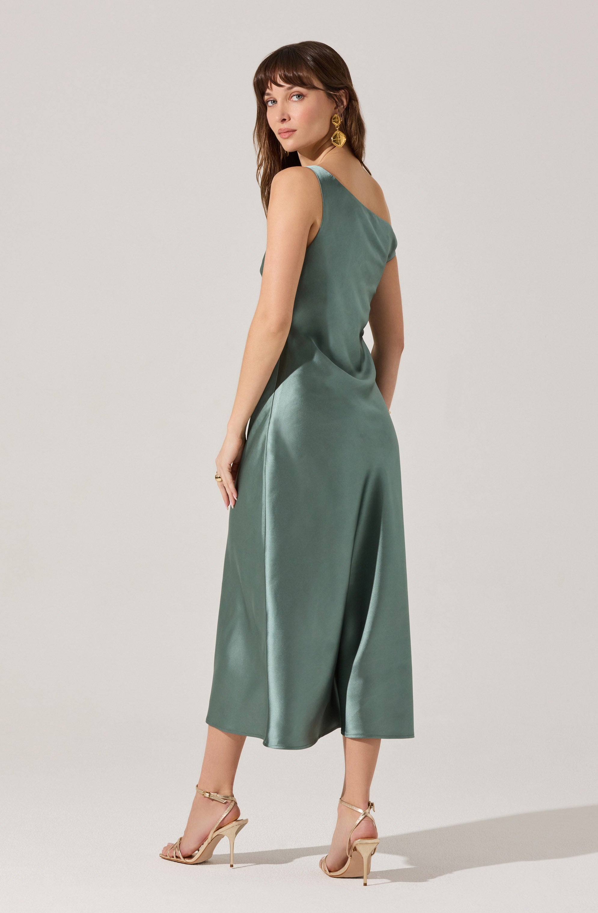 A woman is wearing a sleeveless, satin midi dress in a muted green color. The dress has a smooth, shiny finish and a relaxed fit that drapes elegantly. She pairs the outfit with beige high-heeled sandals and gold statement earrings.